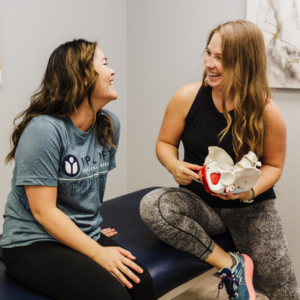 Pelvic floor therapist sitting next to a patient on a treatment table, holding an anatomical model and educating the patient on pelvic floor anatomy.