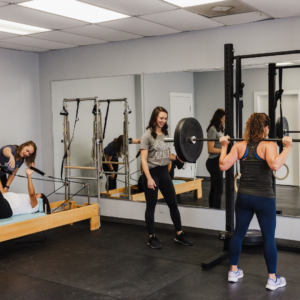 Two pelvic floor therapists assisting patients in a gym setting, with one patient working on core strength on a Pilates reformer and another focusing on squat techniques.