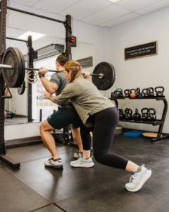 A male patient performs an assisted squat lift while a therapist ensures he is hinging correctly and engaging the right muscles.